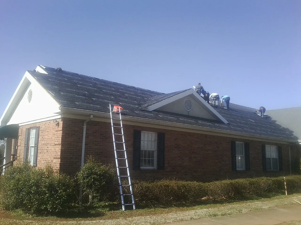 Crew tearing shingles off a brick ranch roof with a ladder set — Emergency Roof Repair work by our crew in Blue Grass
