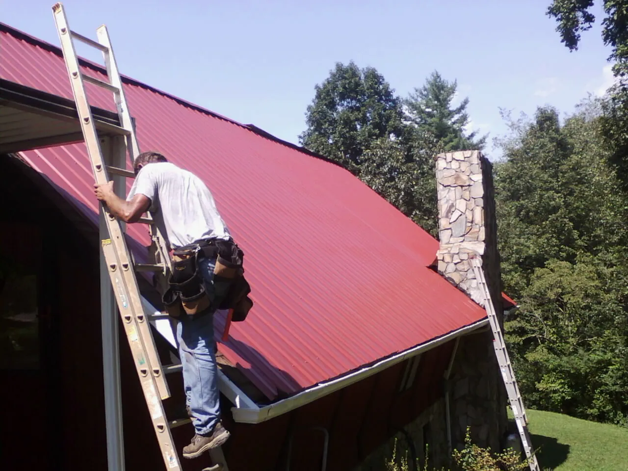 Roofer on a ladder beside a red metal roof and stone chimney — Soffit & Fascia Repair by Roofing Brothers in Blue Grass